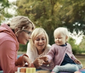 Eine Mutter und ihre Familienpatin spielen auf Augenhöhe mit einem Kleinkind auf einer Picknikdecke.