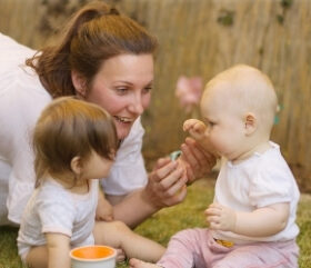 Junge Frau kniet sich auf Augenhöhe zu zwei sitzenden Babys. Sie lächelt die Kinder an.