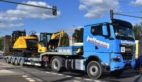 Blauer LKW mit geladenem gelben Bagger der Firma Hoffmann. 