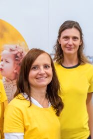 Zwei Frauen in gelben Shirts lächeln sympatisch in die Kamera.